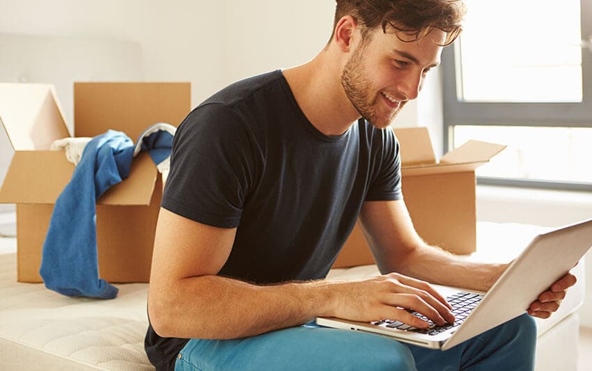 The image features a white hire van parked on a paved residential driveway, with the side door partially open revealing the interior. Inside, there are neatly stacked cardboard boxes and plastic waste containers, indicating recent removal or clearance activity. The van's surface is clean, with a smooth finish and visible signage of Hire Man and Van on the side panel, including subtle green and blue detailing. The surrounding environment includes a grey concrete driveway bordered by a small grassy patch on the right and a tidy front garden with a low brick wall at the rear. Behind the van, there is a suburban residential street with other similar houses and parked vehicles visible in the background, suggesting a typical Maidstone neighbourhood. The lighting is natural daylight, slightly overcast, providing soft, even illumination across the scene. The image captures a moment in a professional rubbish removal process, with the van positioned for efficient loading or unloading of household or garden waste, consistent with local waste management services in the Maidstone area, specifically within postcode ME2.